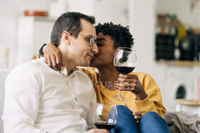 Zufriedenes multiethnisches Paar chillt zu Hause auf dem Sofa mit Rotwein im Glas, während es das Wochenende zu Hause genießt und sich küsst — Stockfoto