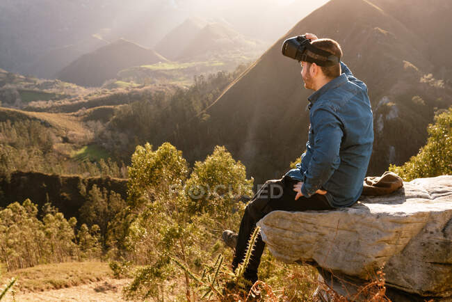 Вид збоку чоловічого мандрівника в окулярах VR, що взаємодіють з віртуальною реальністю, сидячи на пагорбі в гірській місцевості на заході сонця — стокове фото