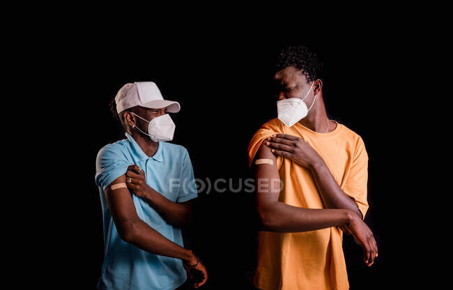 Amigos afro-americanos com rostos máscara protetora com bandagem adesiva gesso no braço depois de obter a vacinação de pé juntos em fundo preto em uma clínica durante o surto de coronavírus — Fotografia de Stock