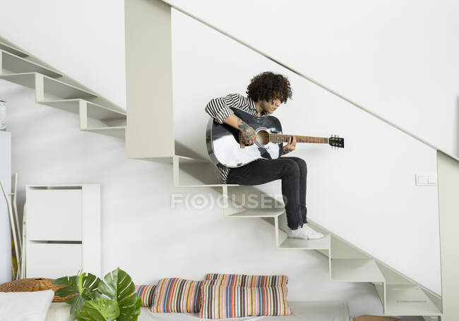 Side view of young hipster male musician playing acoustic guitar while sitting on staircase and looking down in flat — Stock Photo