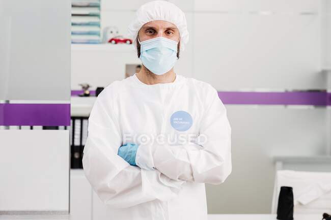 Позитивный врач-мужчина в латексных перчатках и защитной медицинской маске с привитым стикером на белой униформе, стоящий со скрещенными руками в современном медицинском кабинете и смотрящий в камеру — стоковое фото