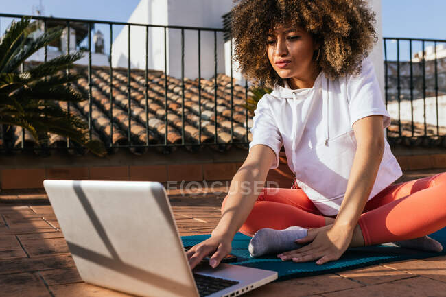 Focada afro-americana fêmea com cabelo encaracolado escolhendo tutorial on-line no laptop enquanto sentado no tapete no telhado e se preparando para a aula de ioga — Fotografia de Stock