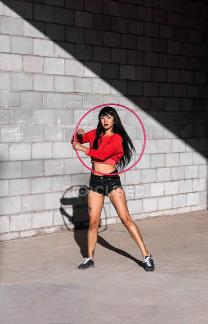 Jeune femme tatouée en vêtements de sport tournoyant hula hoop tout en dansant contre des murs de briques avec des ombres — Photo de stock
