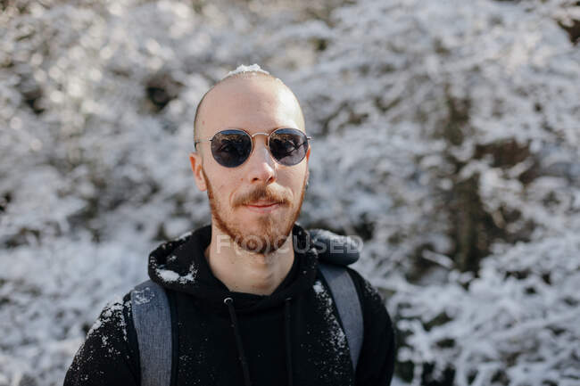 Barbudo mochileiro masculino olhando para a câmera contra árvores nevadas durante a viagem no dia ensolarado — Fotografia de Stock