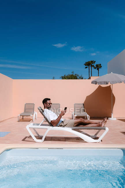 Seitenansicht eines Freiberuflers, der am Pool auf einer Liege liegt und während der Telearbeit im Sommer am sonnigen Tag mit dem Handy im Internet surft — Stockfoto