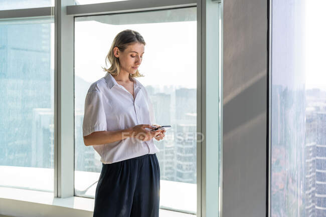 Самотня молода жінка, що стоїть у порожньому офісі з великим вікном, що переглядає мобільний телефон — стокове фото