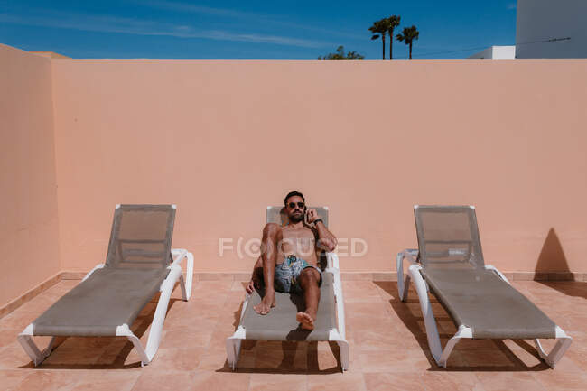 Freelancer masculino enfocado tumbado en una tumbona y tomando el sol mientras discute el proyecto en un smartphone y trabaja remotamente en verano - foto de stock