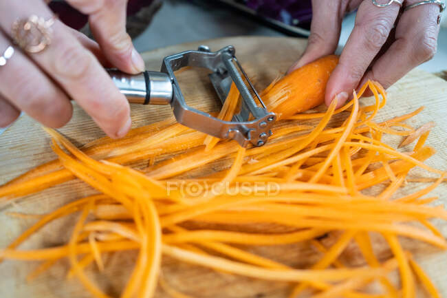 Растениеводство неузнаваемая женщина режет сырую морковь очистителем во время приготовления вегетарианской пищи в доме — стоковое фото