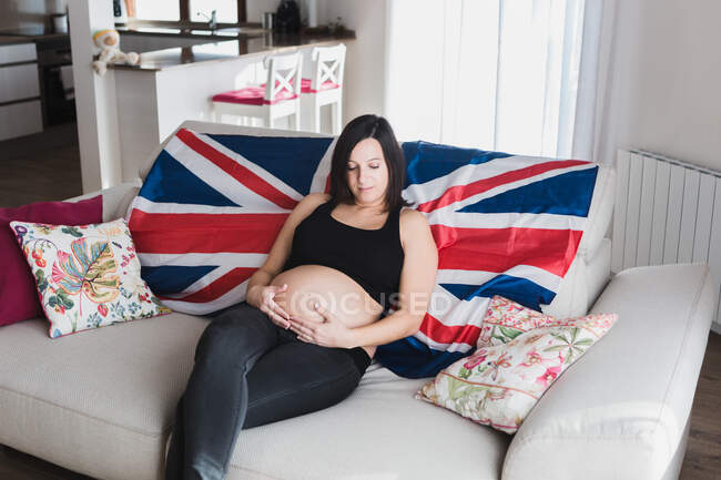 Високий кут делікатного вагітної жінки сидить на дивані і зворушливо животик, насолоджуючись вихідними вдома — стокове фото