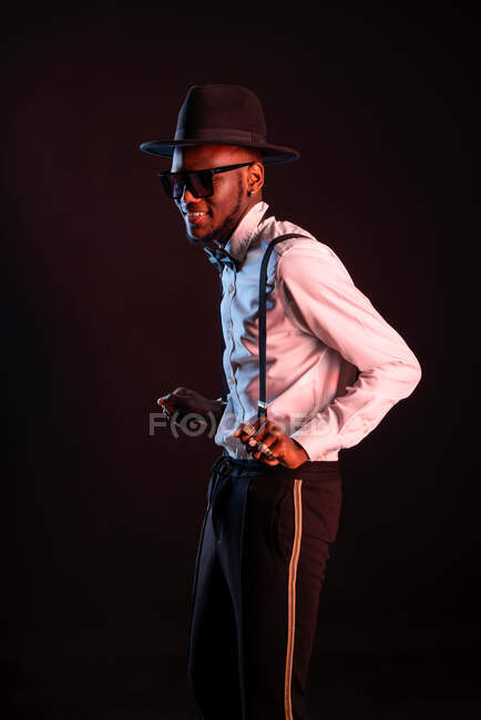 Young masculine ethnic male model in hat and trousers standing dancing while looking away on black background — Stock Photo