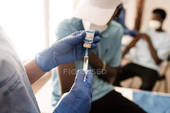 Couper les mains d'un médecin anonyme en gants de latex remplir la seringue du flacon avec le vaccin se préparant à vacciner le patient afro-américain masculin méconnaissable en clinique pendant l'épidémie de coronavirus — Photo de stock