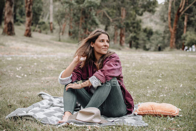 Alegre hembra sentada sobre cuadros en el prado en el bosque y mirando hacia otro lado mientras disfruta de un picnic en Australia - foto de stock