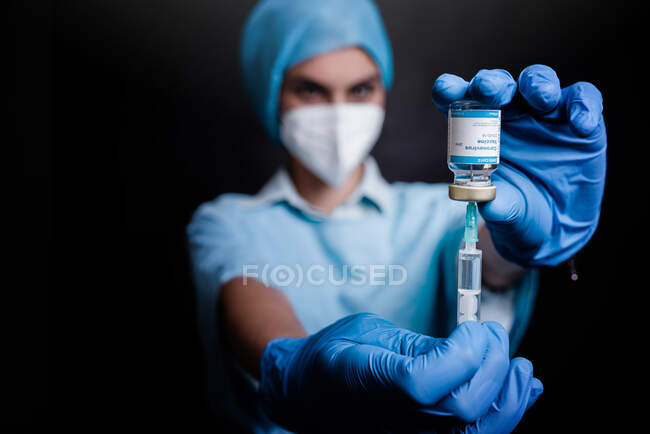 Médico borroso irreconocible en máscara protectora y guantes de látex con vial de vacuna contra el coronavirus y jeringa de pie en fondo negro en la habitación del hospital - foto de stock