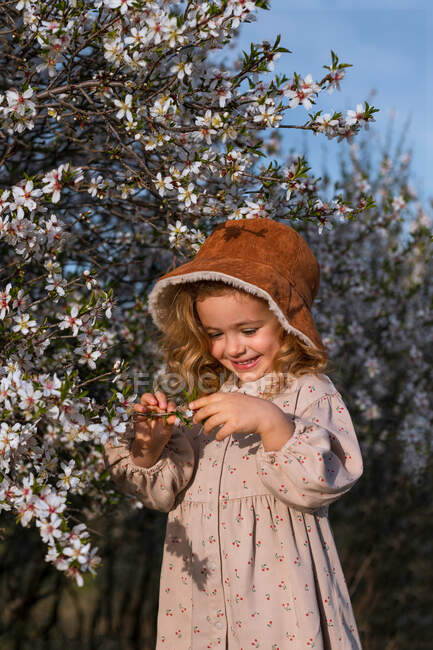 Liebenswert lächelndes kleines Kind im Kleid, das neben blühendem Baum mit Blumen im Frühlingspark steht und nach unten schaut — Stockfoto