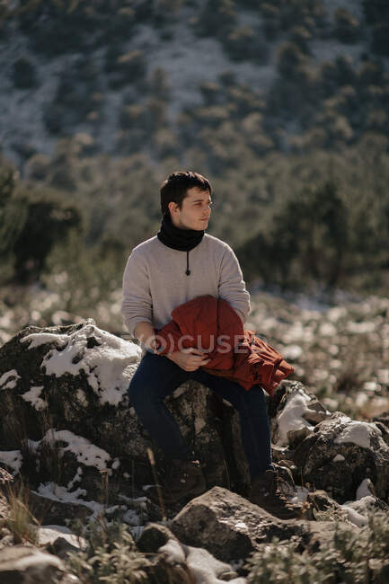 Junge männliche Touristen in warmer Kleidung betrachten die Natur, während sie auf einem Stein gegen den Berg sitzen und im Sonnenlicht wegschauen — Stockfoto