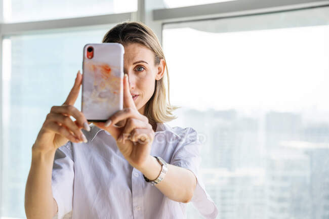 Jeune femme debout dans un bureau vide avec de grandes fenêtres prenant selfie sur le téléphone mobile — Photo de stock