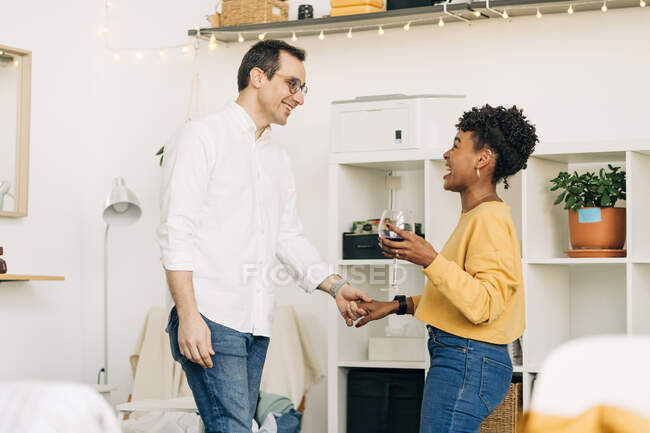 Vista lateral de la sonriente pareja multiétnica enamorada de pie con una copa de vino y cogidas de la mano mientras pasan un fin de semana romántico en casa y se miran mutuamente - foto de stock