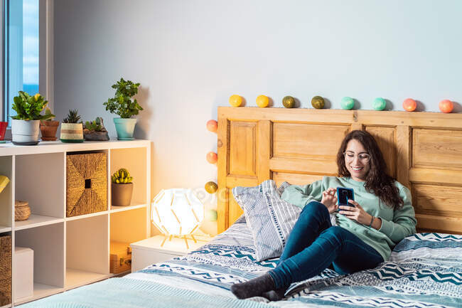 Молодая счастливая женщина в бирюзовой толстовке и очках лежит на кровати с помощью мобильного телефона — стоковое фото