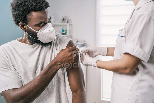 Cultivé spécialiste féminin méconnaissable en uniforme de protection, gants en latex et masque facial vaccinant l'homme afro-américain patient en clinique pendant l'épidémie de coronavirus — Photo de stock