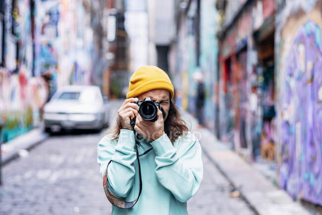 Young hipster male in knitted hat taking photo of graffiti wall on camera in city — Stock Photo