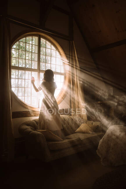 Вид ззаду на невпізнавану жінку в сукні, що стоїть на дивані на круглому вікні в будинку в сонячний день — стокове фото