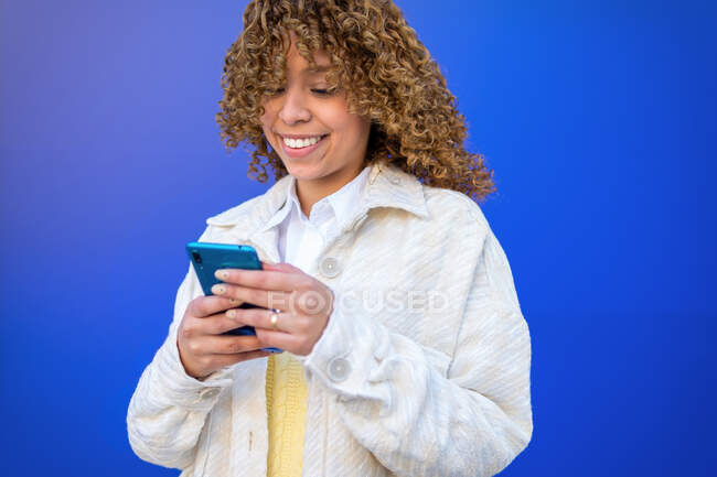Delighted African American female using smartphone while standing on blue background — Stock Photo