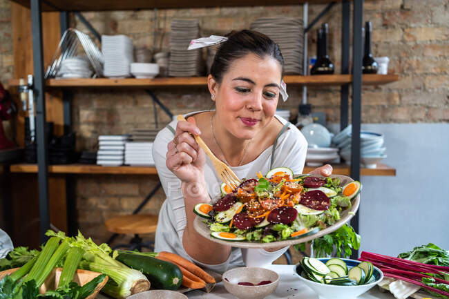 Adult content female with delicious vegetable salad on plate and wooden fork at table against brick wall — Stock Photo