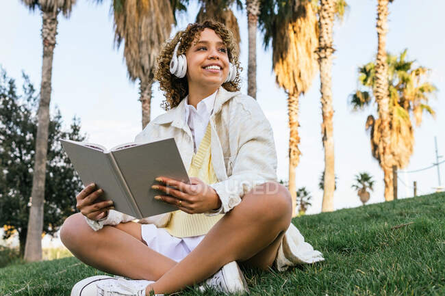 Baixo ângulo de feliz afro-americano fêmea em fones de ouvido sentado no parque exótico e lendo livro interessante no verão — Fotografia de Stock