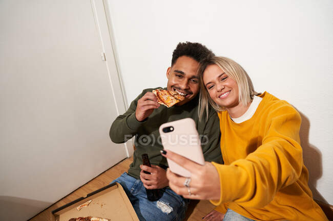 Зверху позитивна багаторасова пара їсть смачну піцу і знімає себе на смартфон, коли охолоджує разом вдома — стокове фото