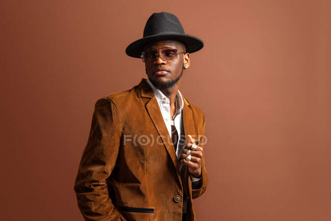 Joven hombre afroamericano en ropa de moda y sombrero mirando hacia otro lado sobre fondo marrón - foto de stock