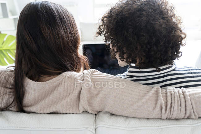 Назад жінка з нетбуком розмовляє з хлопцем, дивлячись на екран на дивані в кімнаті — стокове фото