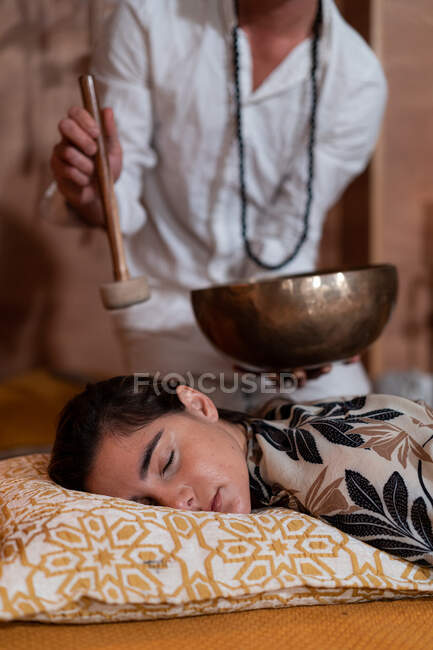 Cortado terapeuta espiritual masculino irreconhecível em miçangas jogando tigela cantando tibetano sobre jovem fêmea com olhos fechados — Fotografia de Stock