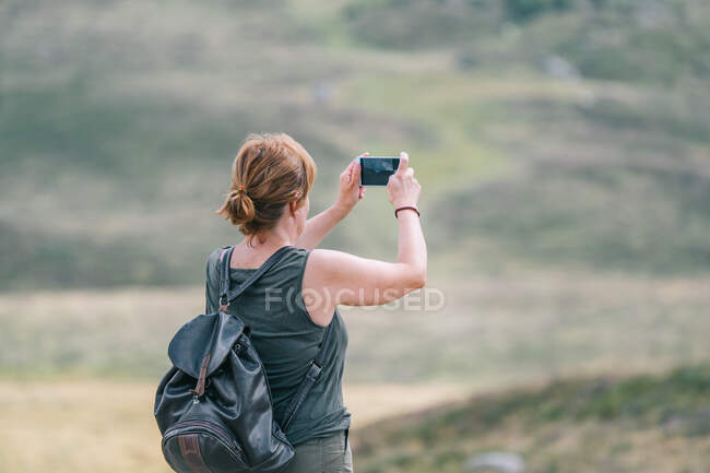 Visão traseira do mochileiro fêmea anônimo tirando fotos de colinas no celular durante a viagem de verão à luz do sol — Fotografia de Stock