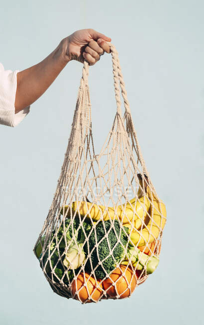 Cultivé femelle méconnaissable debout avec assortiment de fruits et légumes dans un sac de maille écologique contre le mur bleu en ville — Photo de stock