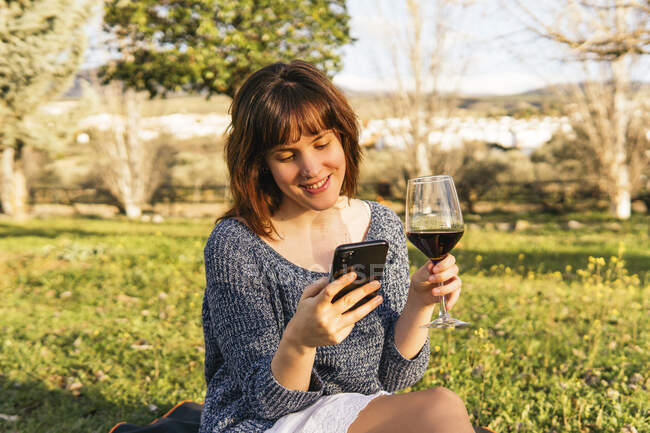 Carefree female sitting on plaid on meadow and using smartphone while enjoying picnic on sunny day in spring — Stock Photo