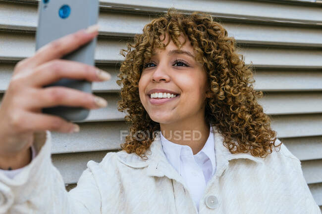 Optimistic African American female with Afro hairstyle taking self portrait on smartphone while standing against metal wall in urban area in city — Stock Photo