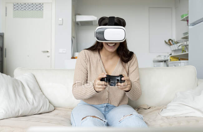 Невпізнавана весела жінка з геймпадком відчуває віртуальну реальність в окулярах, граючи в відеогру на дивані в будинку — стокове фото