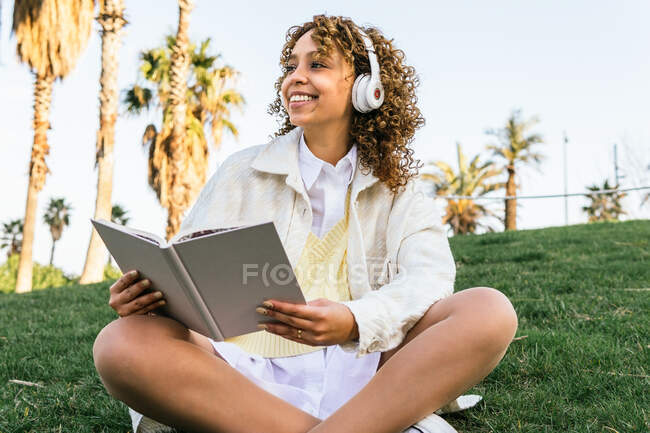 Низкий угол радостной афроамериканки в наушниках, сидящей в экзотическом парке и читающей интересную книгу летом — стоковое фото