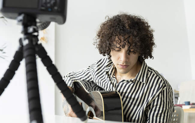 Jovem tatuado guitarrista masculino tocando guitarra acústica durante a gravação de vídeo na câmera de fotos na sala da casa — Fotografia de Stock
