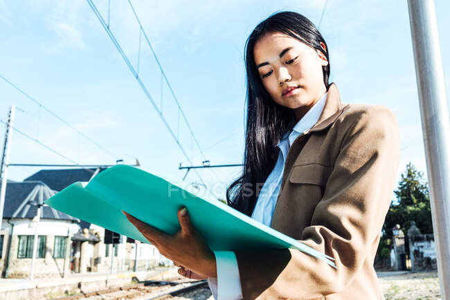 From below focused Asian female entrepreneur reading documents while at railway station and waiting for departure — Stock Photo
