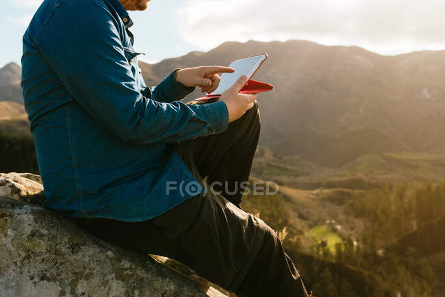 Vista lateral de recortado explorador masculino tranquilo irreconocible con tableta sentada en la roca y disfrutando de una vista espectacular de las montañas en el día soleado - foto de stock