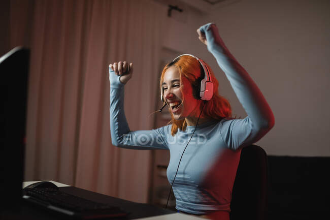 Задоволена жінка-гравець в навушниках, що святкує перемогу в відеоіграх з кулаками вгору, сидячи за столом вдома — стокове фото