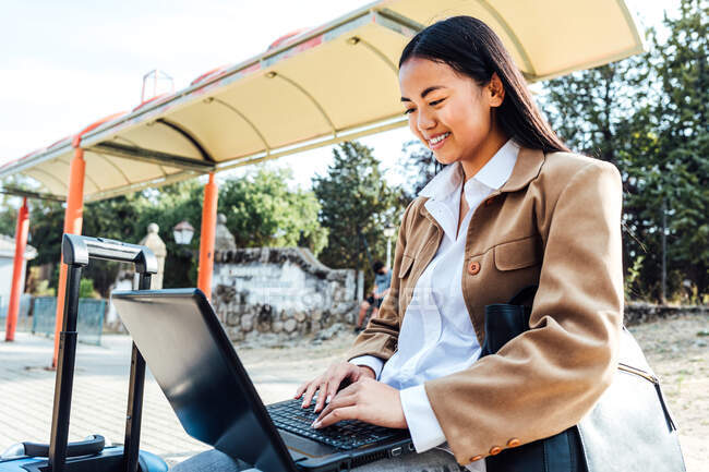 Asian female in mask sitting on bench at railway station and browsing laptop while waiting for train — Stock Photo
