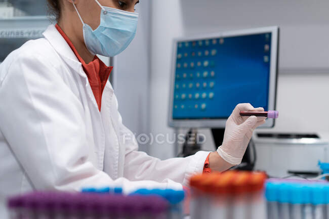Visão lateral de médica fêmea irreconhecível em máscara e luvas segurando tubo com amostra de sangue no laboratório moderno na clínica — Fotografia de Stock