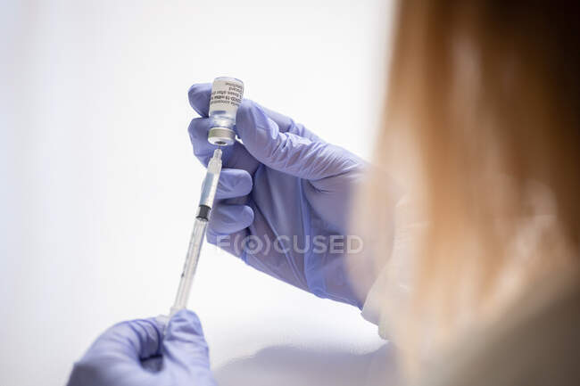 Unrecognizable medic in protective face shield mask and latex gloves with vial of coronavirus vaccine and syringe showing to camera while standing in hospital room — Stock Photo