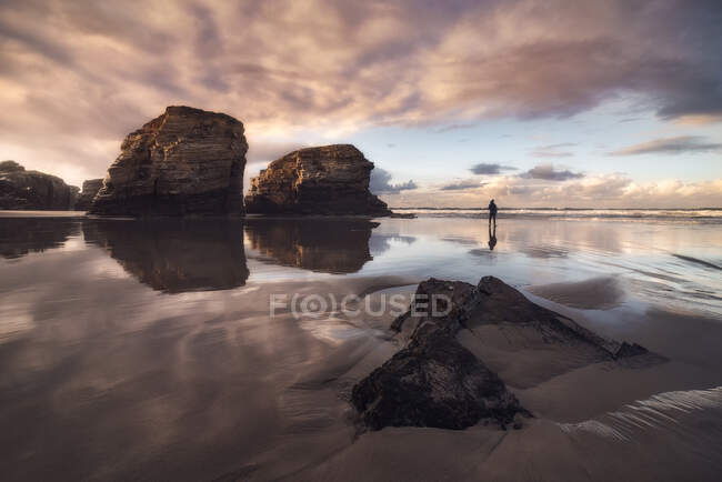 Silhouette de personne anonyme debout sur la plage humide As Catedrais avec des rochers sous le ciel couchant en Galice — Photo de stock