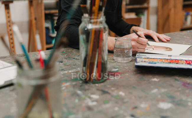 Crop artiste féminine dans la peinture tablier avec aquarelles sur papier tout en étant assis à la table dans l'atelier créatif — Photo de stock