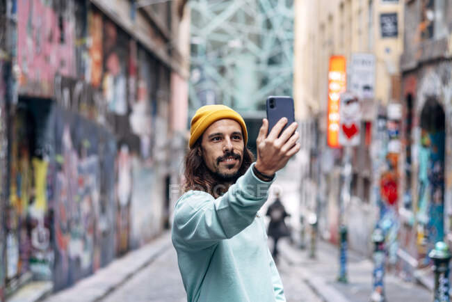 Conteúdo macho com barba em vestuário casual tomando auto retrato no celular na cidade no fundo borrado — Fotografia de Stock