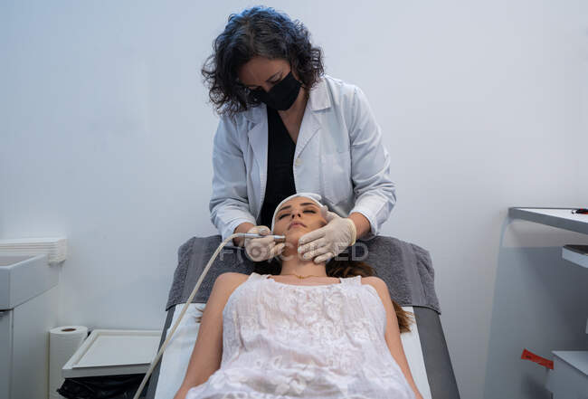 Професійний косметолог використовує спеціальне обладнання і проводить лікування обличчя жінки - клієнтки в сучасній клініці краси. — стокове фото