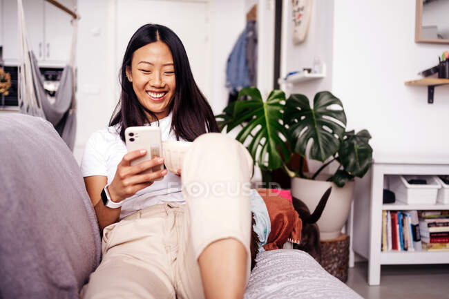 Mensagens de texto feminino étnico alegre no celular enquanto descansa no sofá na sala de estar da casa — Fotografia de Stock
